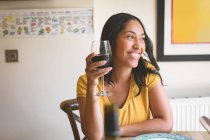 Mujer feliz teniendo vino tinto en la mesa de comedor en casa - foto de stock