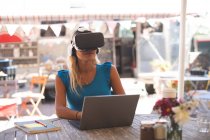Femme utilisant un casque de réalité virtuelle avec ordinateur portable dans un café extérieur — Photo de stock