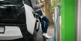 Женщина пьет кофе во время зарядки электромобиля на зарядной станции — стоковое фото