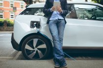 Low section of businessman using digital tablet while charging electric car at charging station — Stock Photo