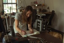 Designer de jóias feminino escrevendo em um registro na oficina — Fotografia de Stock