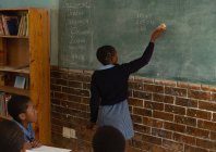 Schoolgirl writing on chalkboard in classroom at school — Stock Photo