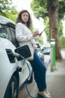 Ділова жінка з чашкою кави заряджає електричний автомобіль на зарядній станції — стокове фото