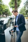 Geschäftsmann nutzt Handy beim Laden von Elektroauto an Ladestation — Stockfoto