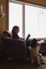 Старшая женщина читает книгу, пока собака сидит рядом с ней в гостиной — стоковое фото