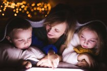 Primer plano de la madre y los niños debajo de la manta usando tableta digital contra las luces de Navidad - foto de stock
