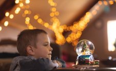 Curieux garçon regardant boule de neige arbre de Noël à la maison — Photo de stock