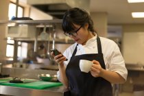 Жіночий шеф-кухар використовує мобільний телефон, маючи каву на кухні — стокове фото