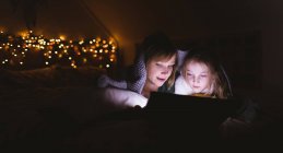 Mãe e filha sob o cobertor usando tablet digital contra luzes de Natal — Fotografia de Stock