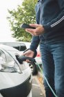 Seção média do homem usando o telefone móvel ao carregar o carro elétrico na estação de carregamento — Fotografia de Stock