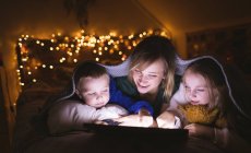 Мати і діти під ковдрою використовують цифровий планшет проти різдвяних вогнів — стокове фото