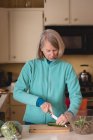 Seniorin schneidet Gurke mit Messer auf Schneidebrett in Küche — Stockfoto