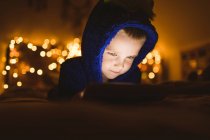 Крупный план мальчика в синей куртке с помощью цифрового планшета против рождественских огней — стоковое фото