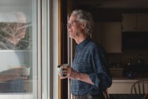 Старший чоловік дивиться крізь вікно, маючи чашку кави вдома — стокове фото