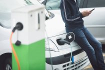 Partie médiane de l'homme utilisant un téléphone portable tout en rechargeant la voiture électrique à la station de charge — Photo de stock