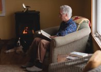 Старший чоловік читає книгу на кріслі для руки у вітальні вдома — стокове фото