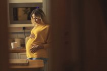 Беременная женщина прикасается к животу на кухне дома — стоковое фото