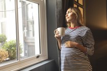 Беременная женщина с кофейной чашкой стоит у окна дома — стоковое фото
