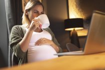Mulher grávida bebendo café enquanto usa laptop em casa — Fotografia de Stock