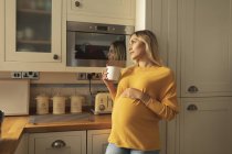 Задумчивая беременная женщина с чашкой кофе, стоящей на кухне — стоковое фото
