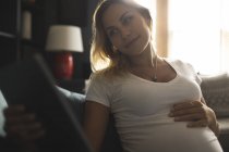 Pregnant woman using digital tablet on sofa at home — Stock Photo