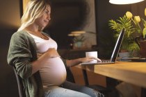Schwangere berührt ihren Bauch und benutzt Laptop zu Hause — Stockfoto