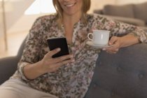 Media sezione di donna che utilizza il telefono cellulare mentre prende il caffè a casa — Foto stock