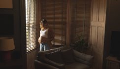 Беременная женщина прикасается к животу у окна дома — стоковое фото