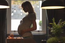 Pregnant woman standing next to the window touching her belly at home — Stock Photo