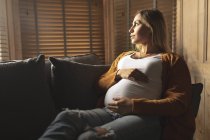 Mulher grávida sentada no sofá olhando através da janela em casa — Fotografia de Stock