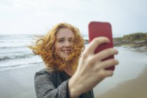 Donna rossa scattare selfie con il telefono cellulare in spiaggia . — Foto stock