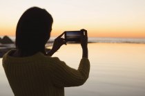 Woman taking photo with mobile phone on beach during sunset — Stock Photo