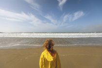 Visão traseira da mulher ruiva em casaco amarelo em pé na praia . — Fotografia de Stock