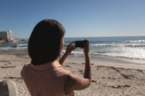 Вид ззаду жінки, фотографується з мобільним телефоном біля пляжу — стокове фото