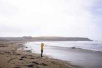Vista lateral da mulher de casaco amarelo em pé na praia . — Fotografia de Stock