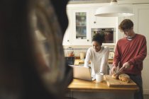 Frau benutzt Laptop, während Mann in Küche zu Hause Laib Brot schneidet — Stockfoto