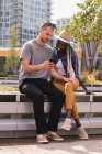 Улыбающаяся пара, сидящая на скамейке и пользующаяся мобильным телефоном в городе — стоковое фото