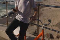 Середина чоловіка з велосипедом, використовуючи мобільний телефон біля перил на пляжі — стокове фото
