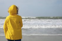 Visão traseira da mulher de jaqueta amarela em pé na praia . — Fotografia de Stock