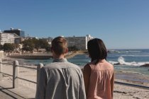 Vista trasera de la pareja caminando en el paseo marítimo cerca de la playa - foto de stock