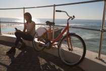 Mujer con bicicleta usando teléfono móvil en el paseo marítimo cerca de la playa - foto de stock