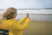 Рыжая женщина фотографирует с мобильного телефона на пляже . — стоковое фото