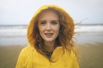 Portrait of redhead woman in yellow jacket standing in beach. — Stock Photo