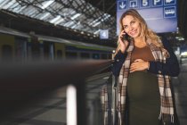 Щаслива вагітна жінка розмовляє по мобільному телефону на вокзалі — стокове фото