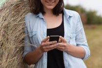 Средняя часть женщины, которая пользуется мобильным телефоном, опираясь на тюк сена — стоковое фото