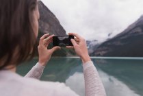 Вид ззаду жінки, що клацає фотографіями з мобільним телефоном біля озера — стокове фото