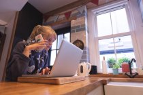 Mulher falando no celular enquanto usa laptop em casa — Fotografia de Stock