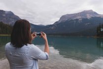 Вид сзади женщины, щёлкающей по фотографиям с мобильного телефона рядом с озером — стоковое фото