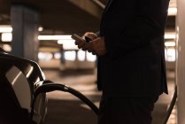 Empresário usando telefone celular ao carregar carro elétrico na estação de carregamento — Fotografia de Stock