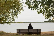Rückansicht einer Frau, die auf einer Bank am Flussufer sitzt — Stockfoto